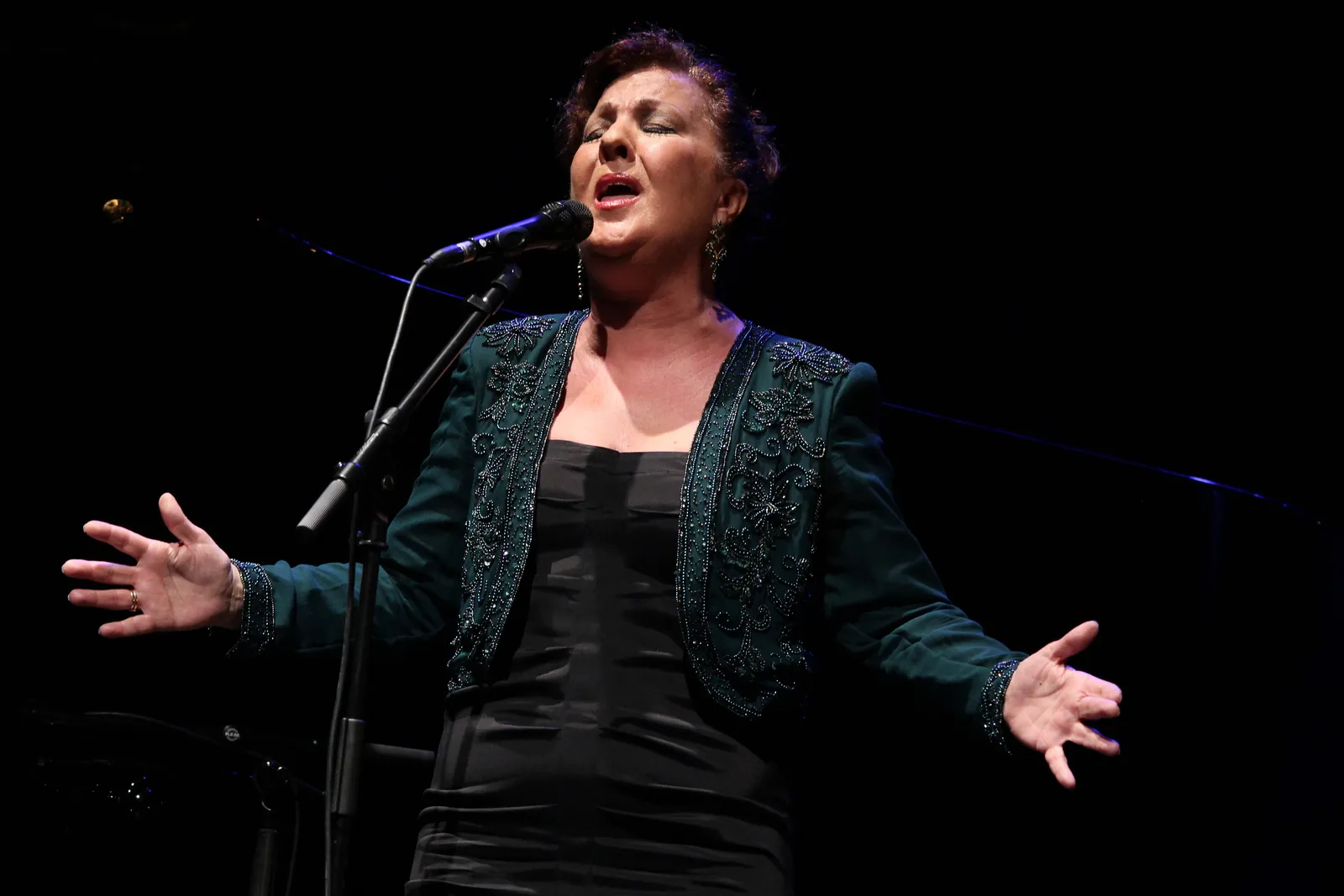 A flamenco singer on stage with her arms open and a microphone in front of her, lit against a dark background