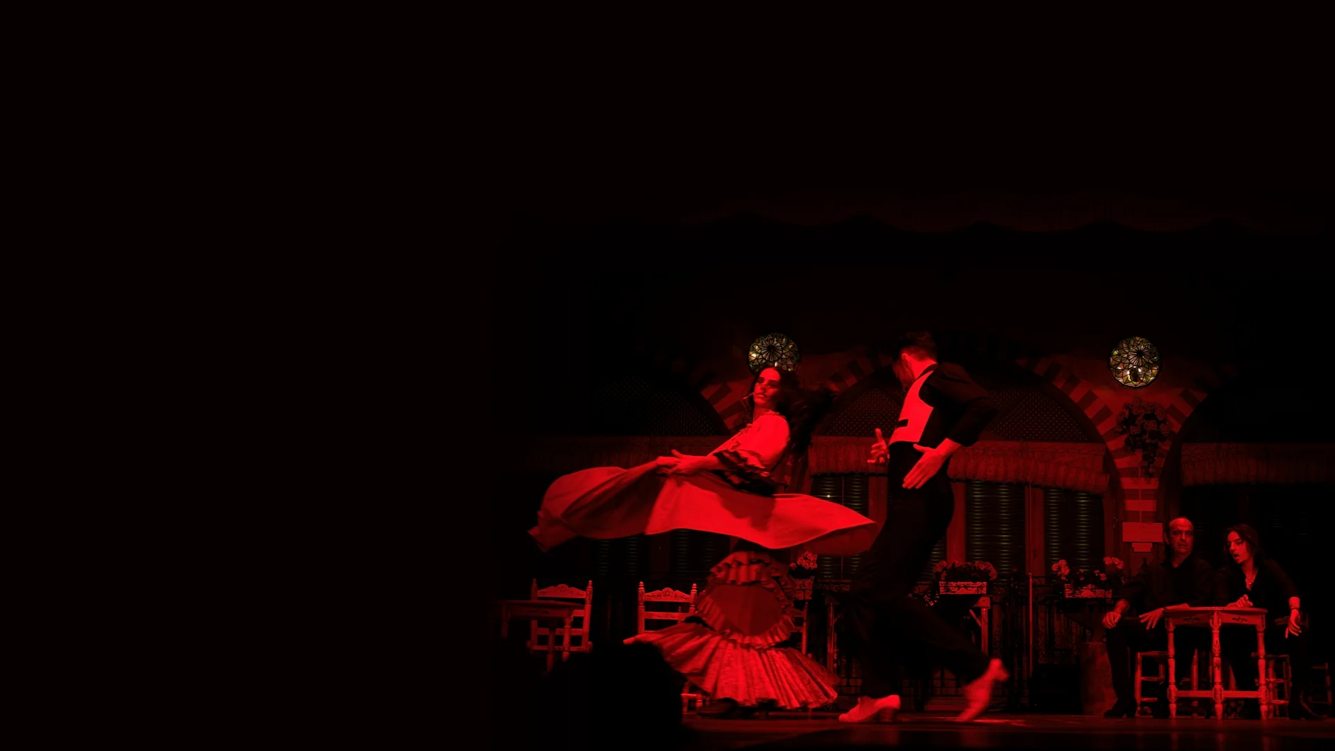 Two flamenco dancers, a woman in a red dress mid-turn and a man in a black waistcoat, on a stage lit in red, with two performers seated at a small table on the right